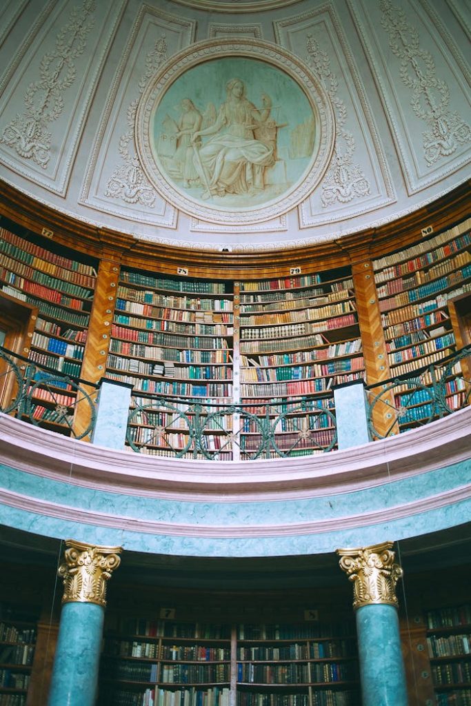 Low angle of assorted literature on bookshelves in aged masonry museum with columns and picture on wall